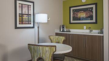 A hotel room with a round dining table, two chairs, a floor lamp, a coffee maker, and a framed picture of a stadium on the wall.