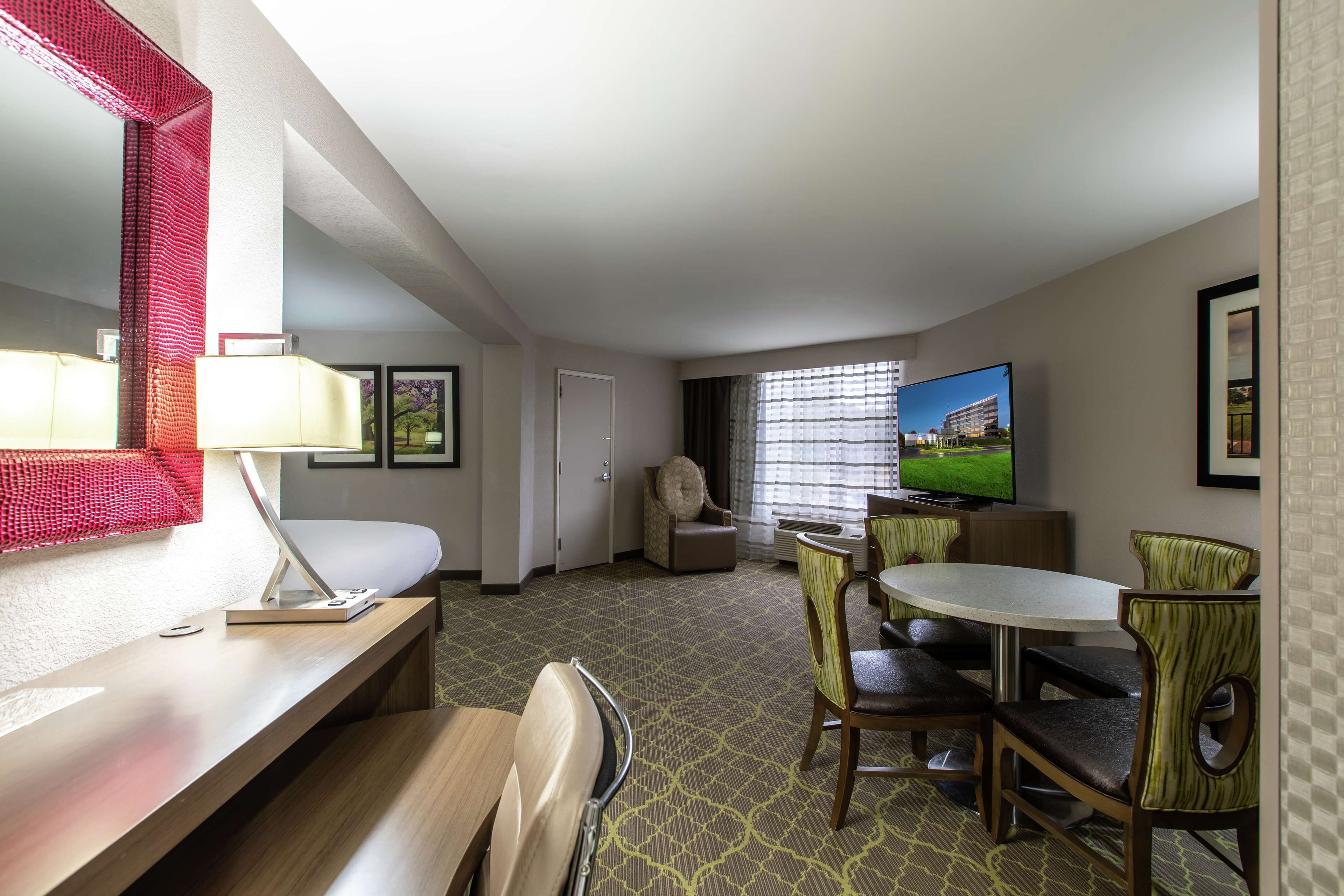 A hotel room with a bed, a desk with a lamp, a TV, a dining area with chairs, and a window with curtains.