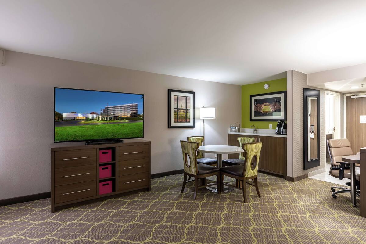 A hotel room with a television, a dining area with chairs, a small kitchen area, and a door leading to another room.