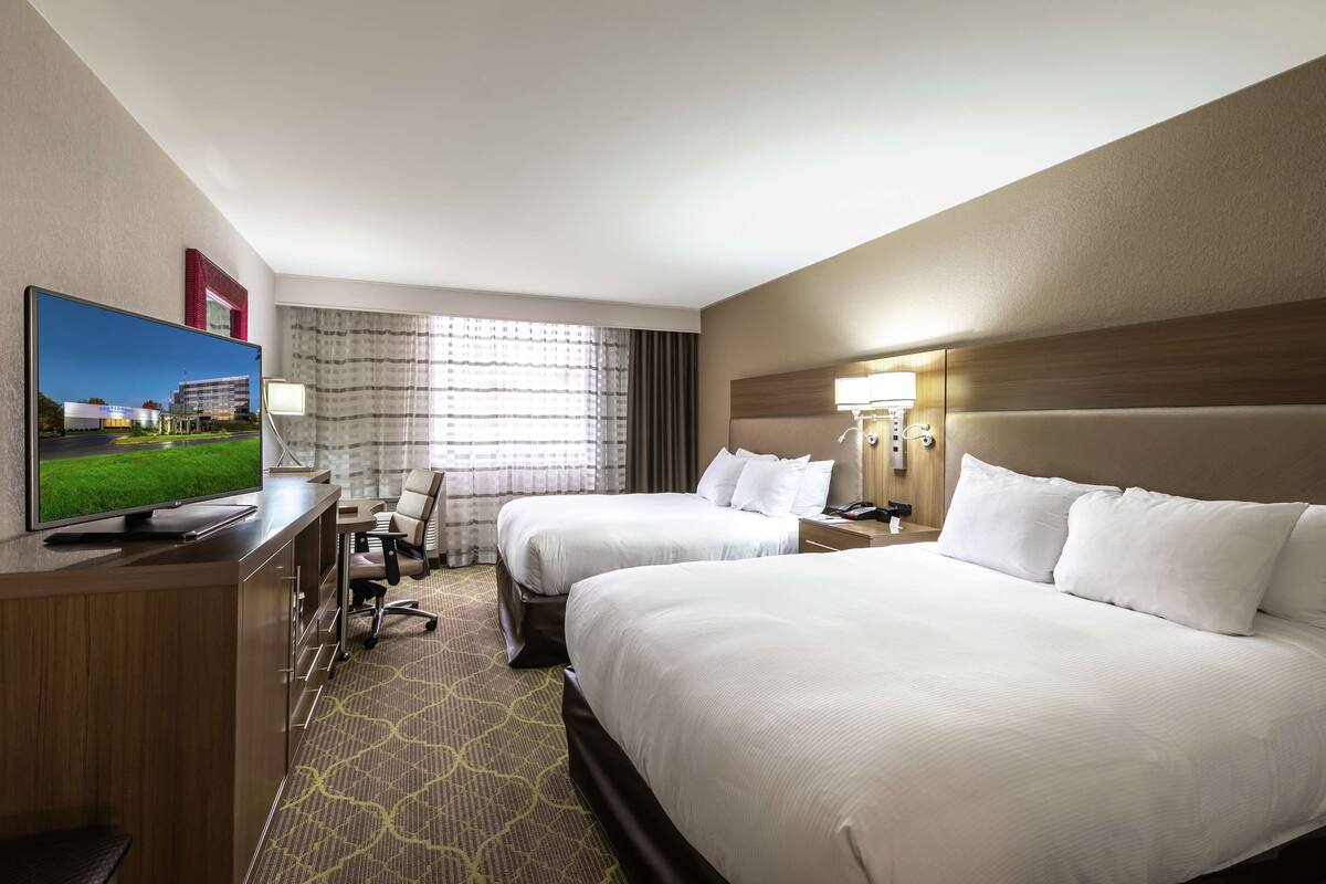A hotel room with two beds, a desk with a flat-screen TV, a chair, a lamp, and a window with curtains.