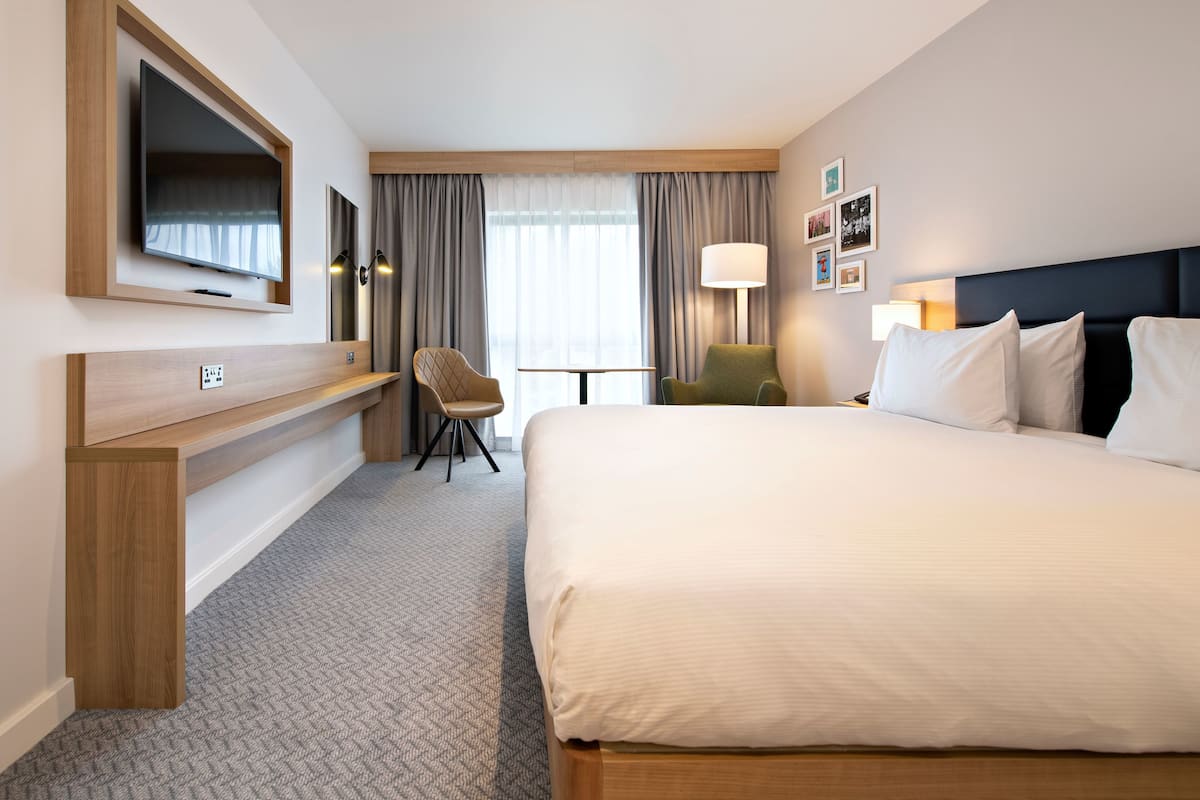A hotel room with a large bed, a desk with a chair, a television mounted on the wall, and a window with curtains.
