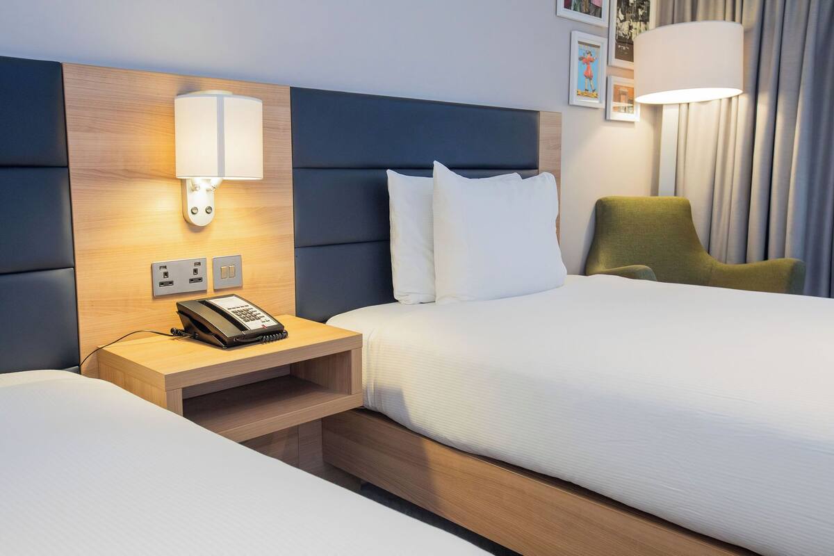 A hotel room with a bed, bedside table with a phone, wall-mounted lamp, and a green chair.