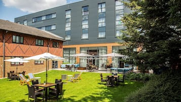 A hotel with a modern exterior, outdoor seating area with umbrellas, and a swimming pool.