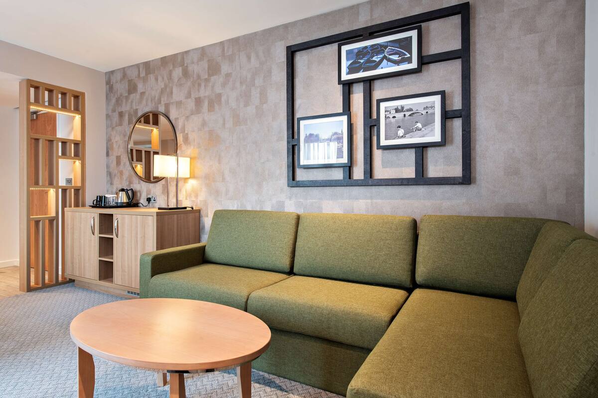 A modern living room with a green sofa, a round wooden coffee table, a wooden cabinet with a mirror, and framed pictures on the wall.
