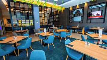 Un restaurante moderno con sillas azules y mesas de madera, una pantalla de televisión en la pared y plantas decorativas.