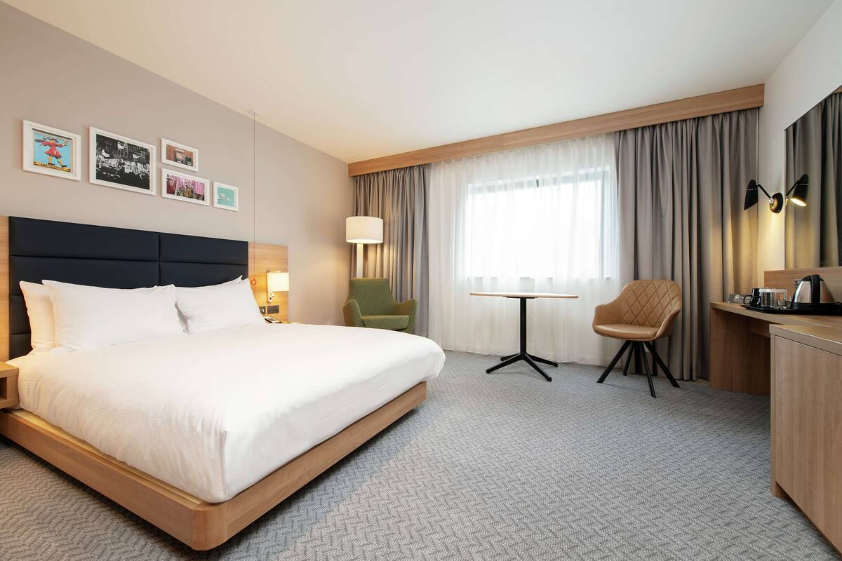 A hotel room with a large bed, a desk, a chair, a lamp, and a window with curtains.