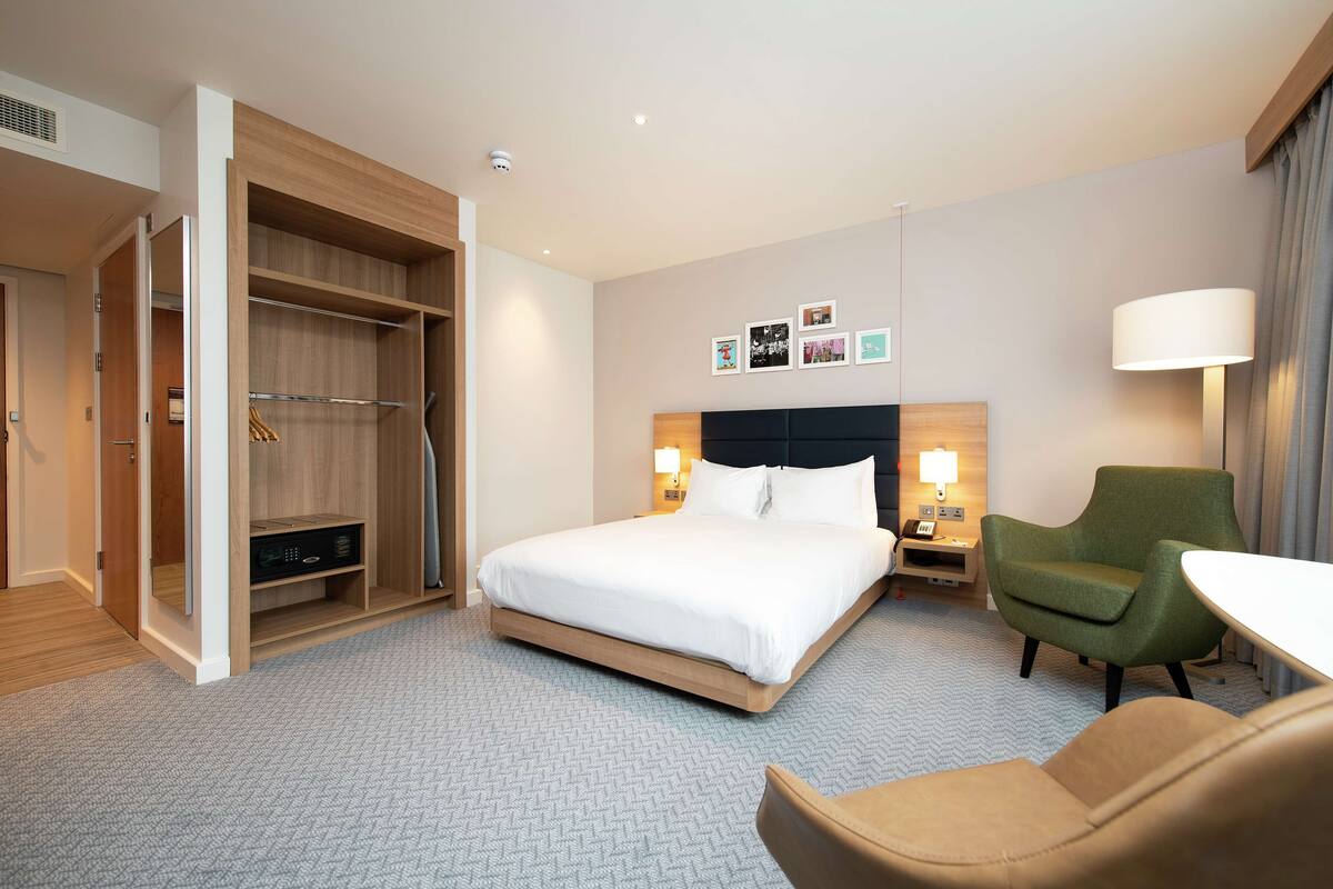 A hotel room with a bed, a green armchair, a brown armchair, a desk, and a wardrobe.