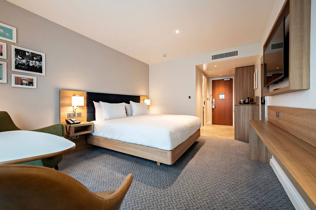 A hotel room with a bed, bedside table, lamp, chair, and a TV mounted on the wall.