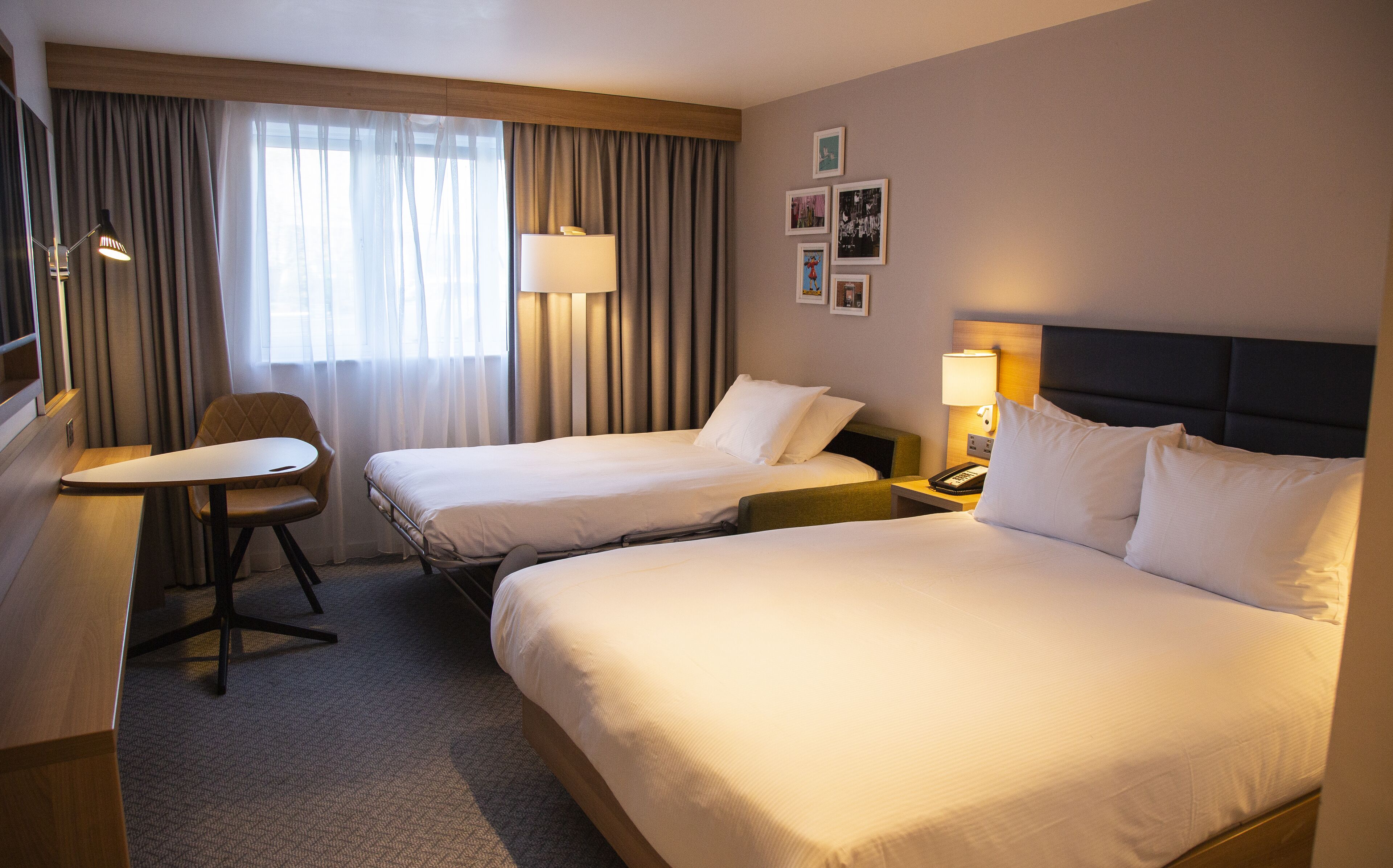 A hotel room with two beds, a desk, a chair, and a nightstand with a lamp.