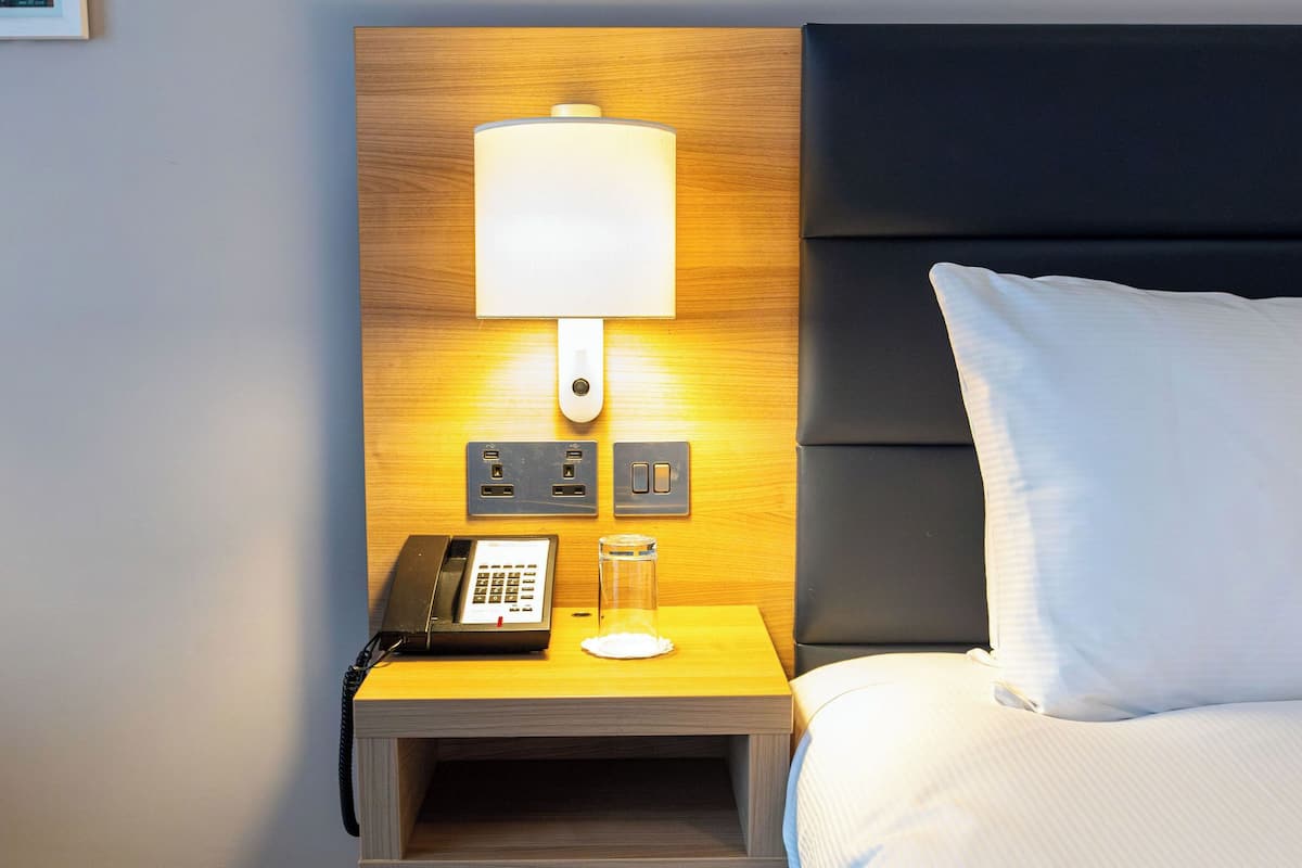 A hotel room with a wooden nightstand, a lamp, a phone, and a glass jar.