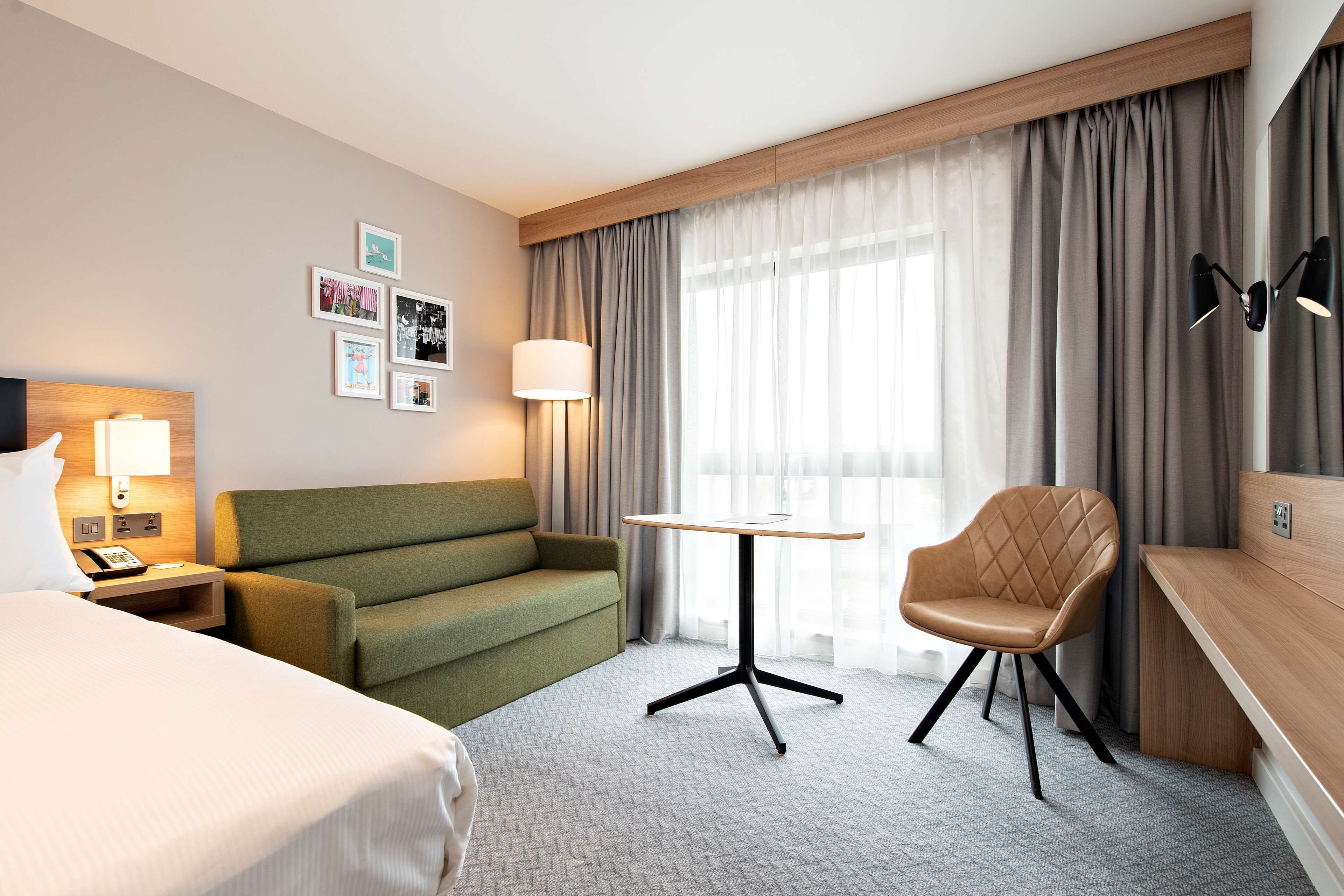 Habitación de hotel con cama, sofá verde, escritorio de madera, silla, lámpara y ventana con cortinas.