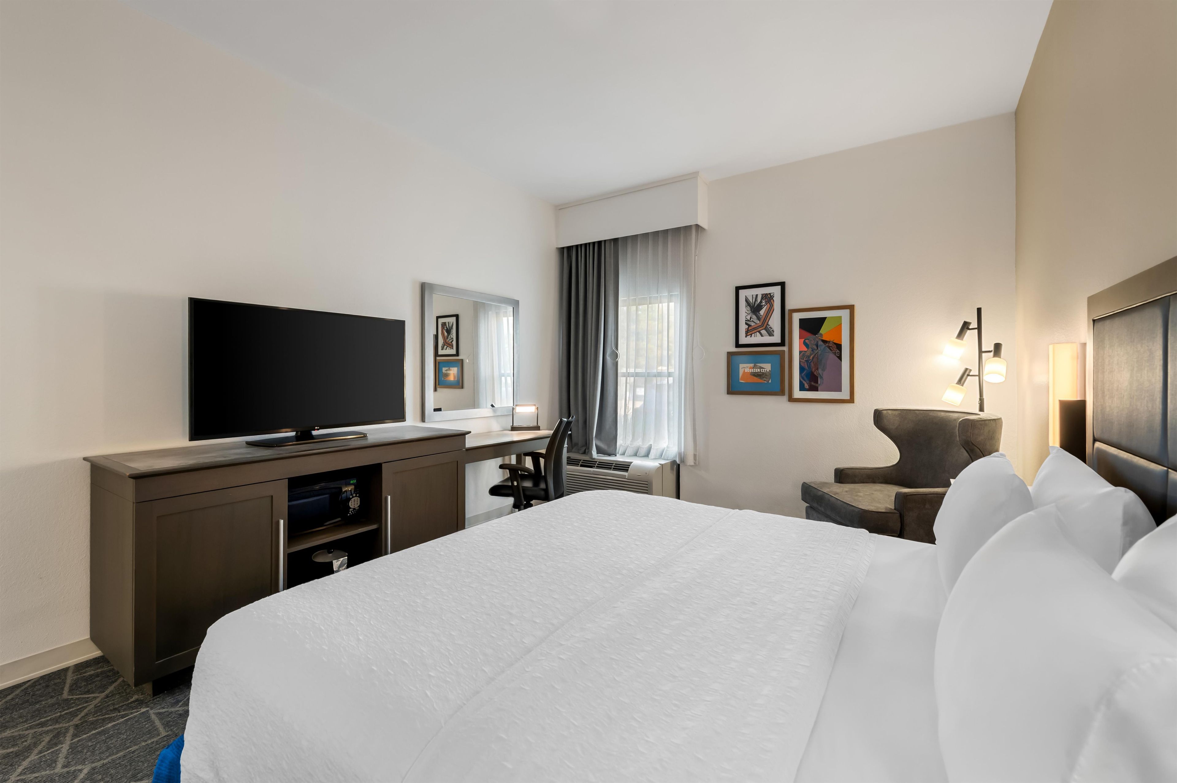 A hotel room with a large bed, a desk with a flat-screen TV, a chair, and a desk lamp.