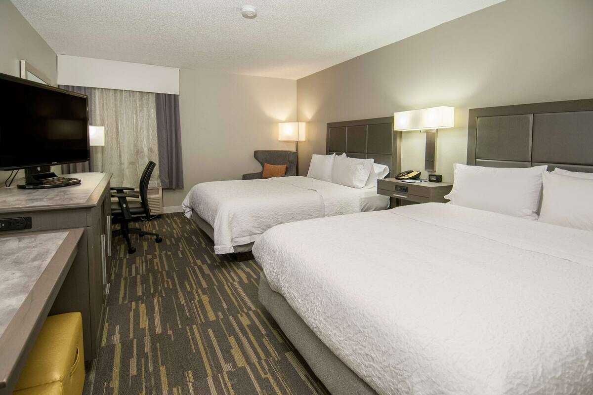 A hotel room with two beds, a desk with a television, a chair, and a lamp.