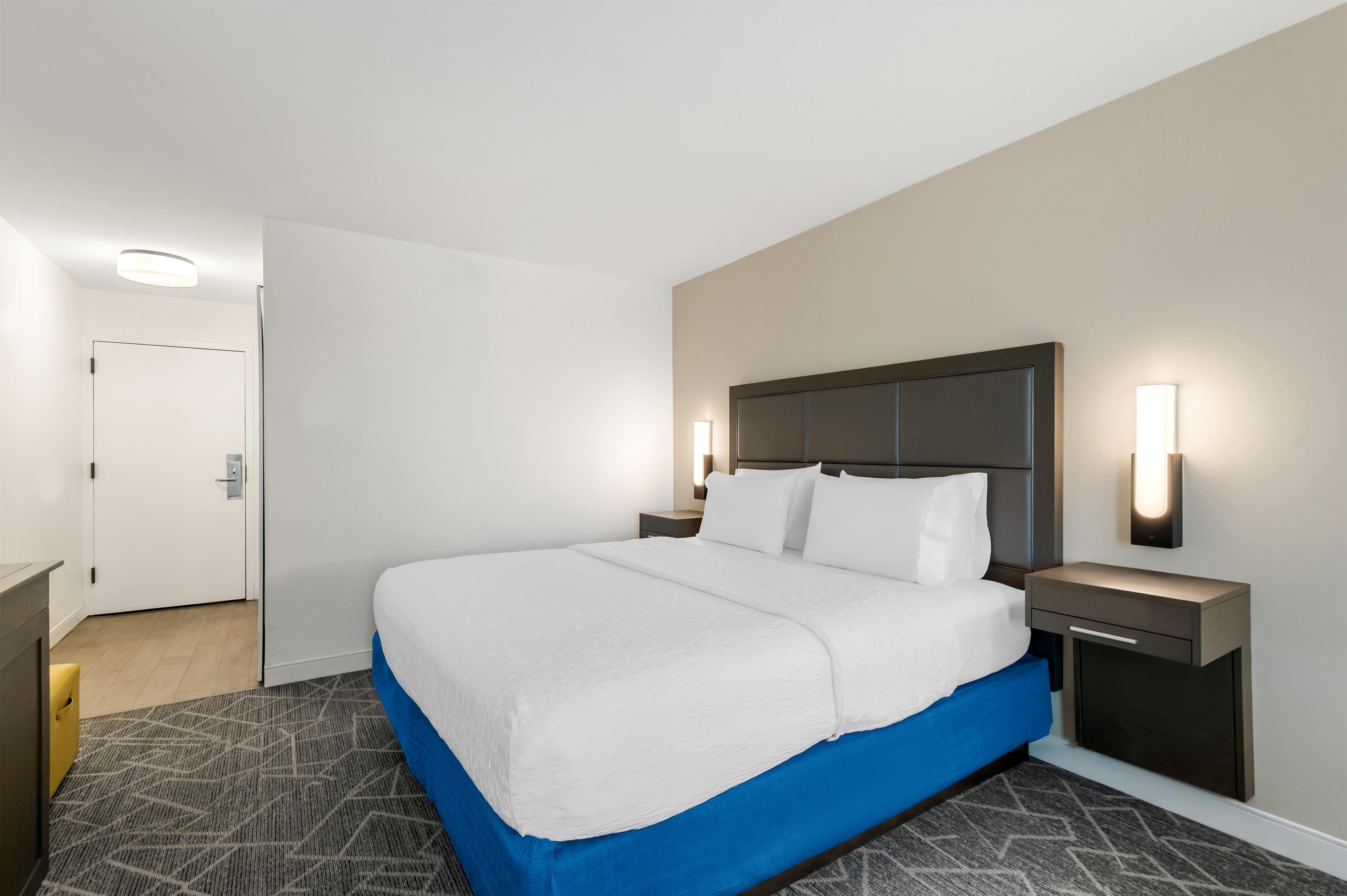 A hotel room with a bed, a nightstand, a wall-mounted lamp, and a door.