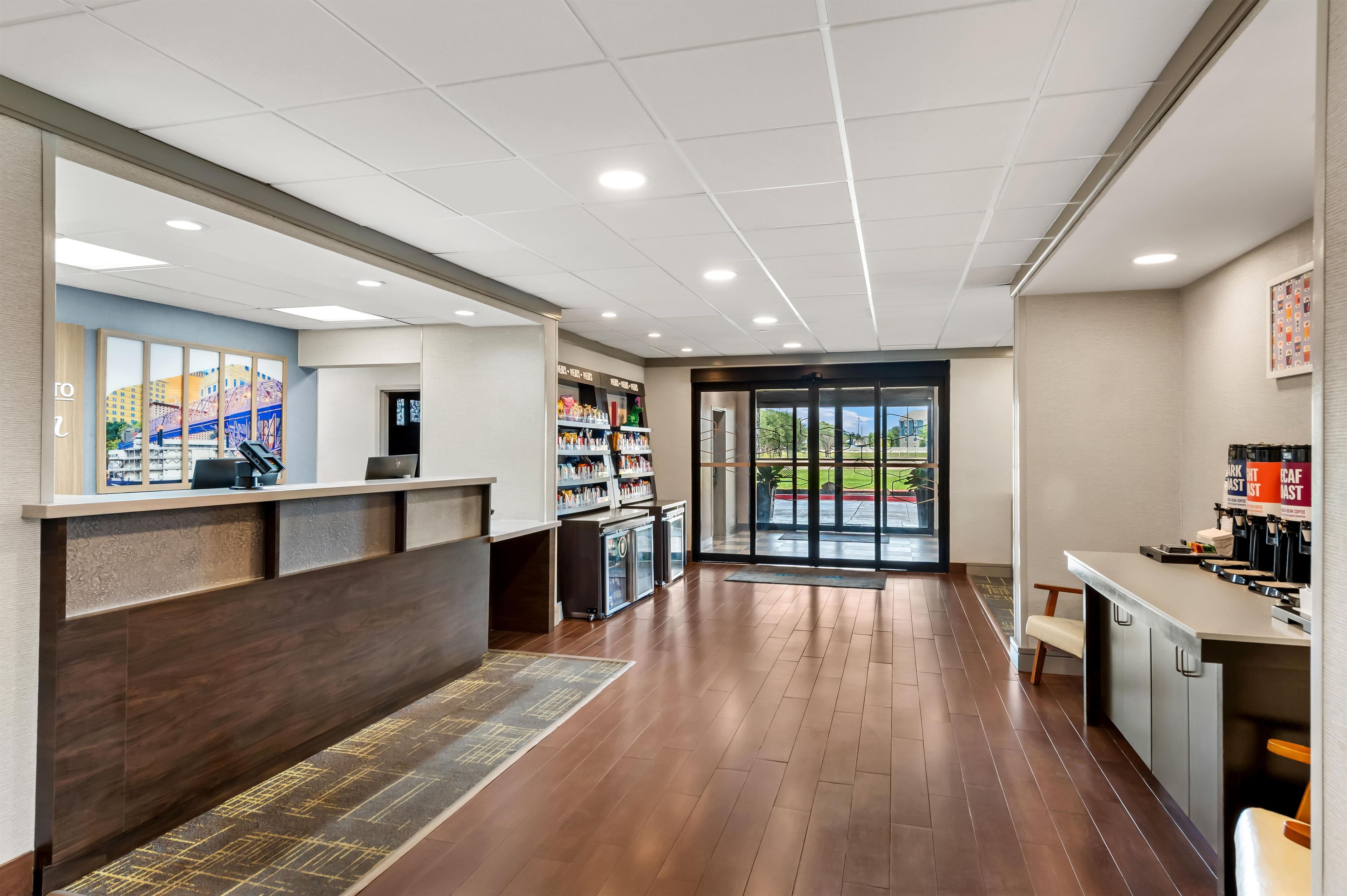 A hotel lobby with a reception desk, a coffee station, and a view through the glass doors.