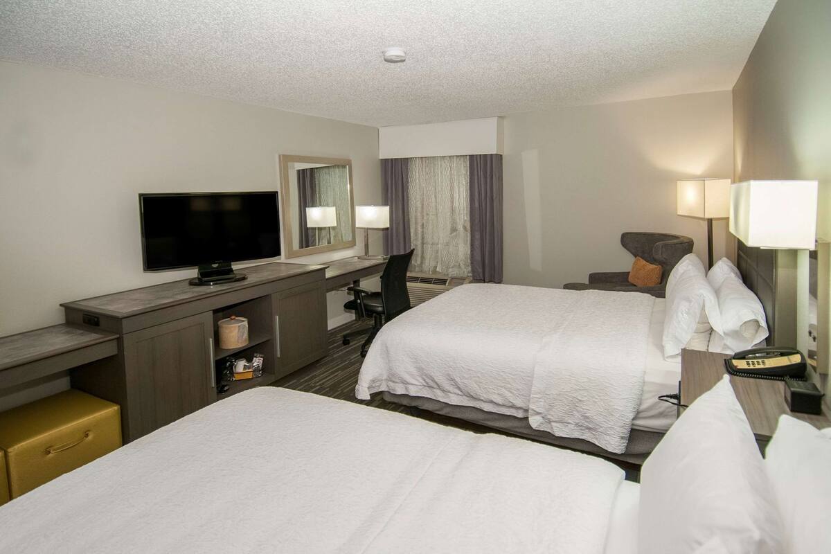 A hotel room with two beds, a desk, a television, a mirror, and a lamp.