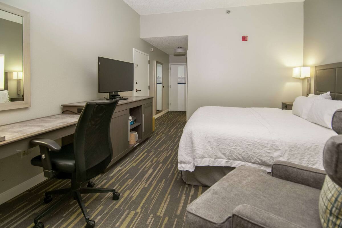A hotel room with a bed, a desk with a chair, a television, and a small table with a lamp.