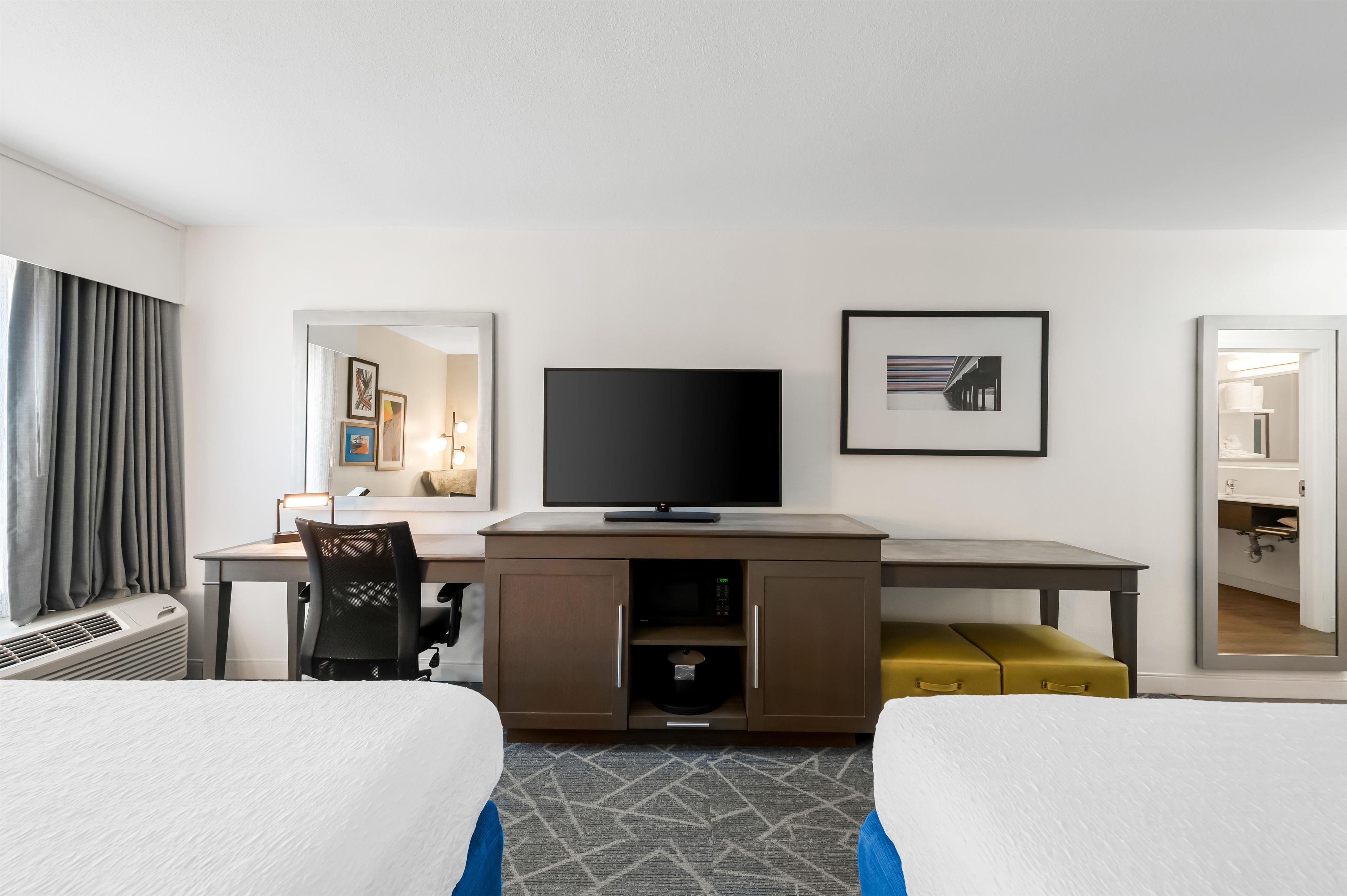 A hotel room with a bed, a desk with a flat-screen TV, a chair, a small sofa, and a mirror.