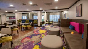 Un hall de hotel con una zona de estar central, varias mesas y sillas. Se observan ventanas grandes con cortinas, un mostrador de recepción y obras de arte en las paredes.