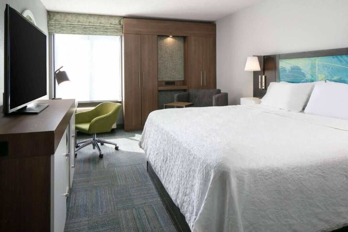 Una habitación de hotel con una cama, un escritorio con televisor, una silla, una mesita de noche y una ventana con cortinas.