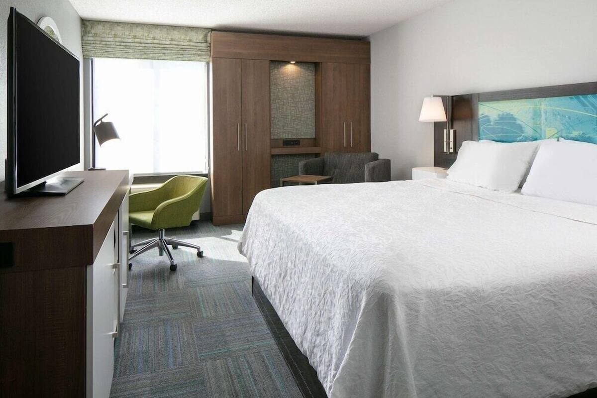 A hotel room with a large bed, a desk with a flat-screen TV, a green chair, a nightstand with a lamp, and a large window with curtains.