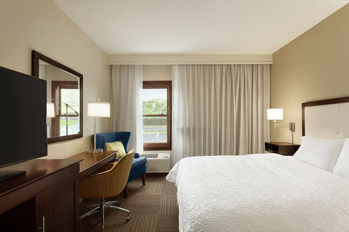 A hotel room with a bed, a desk with a chair, a television, a window with curtains, and a nightstand with a lamp.
