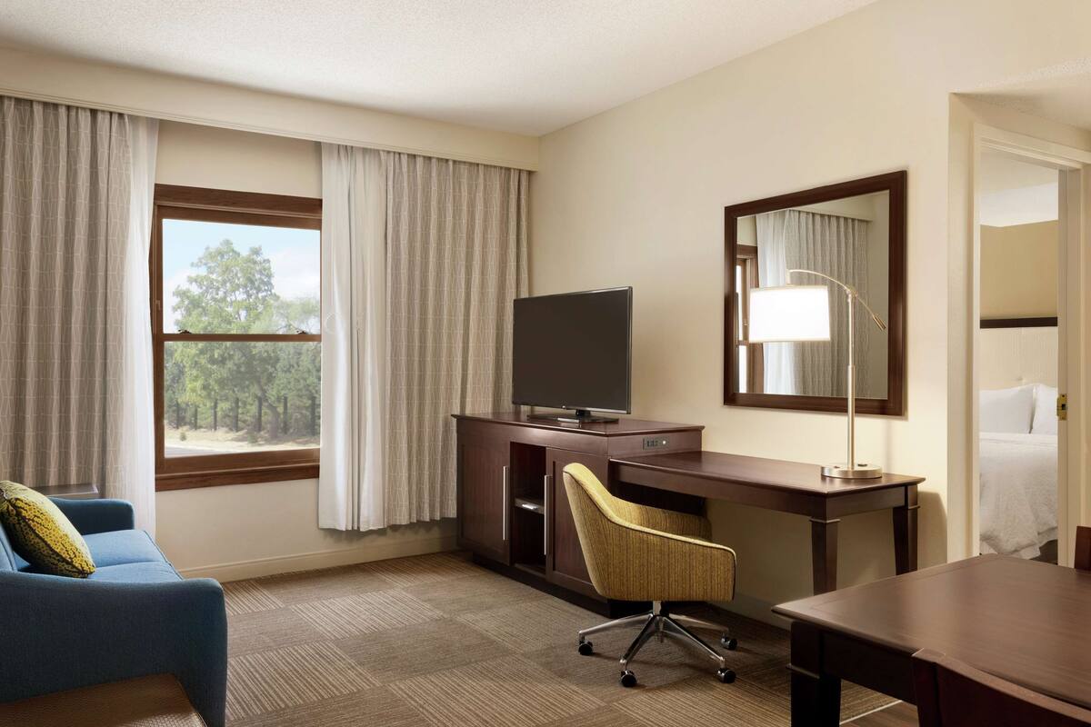 A hotel room with a bed, a desk with a computer, a chair, a TV on a cabinet, and a window with curtains.