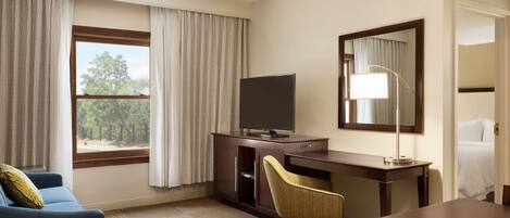 A hotel room with a bed, a desk with a computer, a chair, a TV on a cabinet, and a window with curtains.