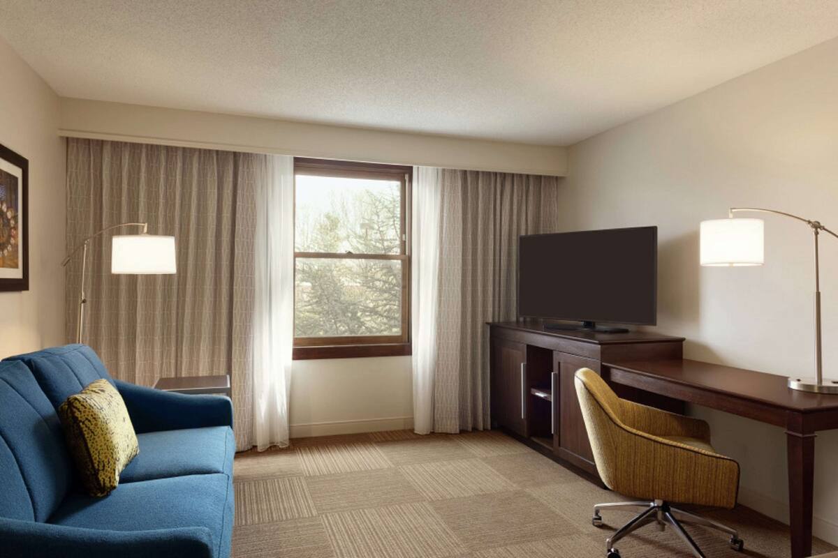 A hotel room with a large window, a flat-screen TV, a desk, a chair, and a sofa.
