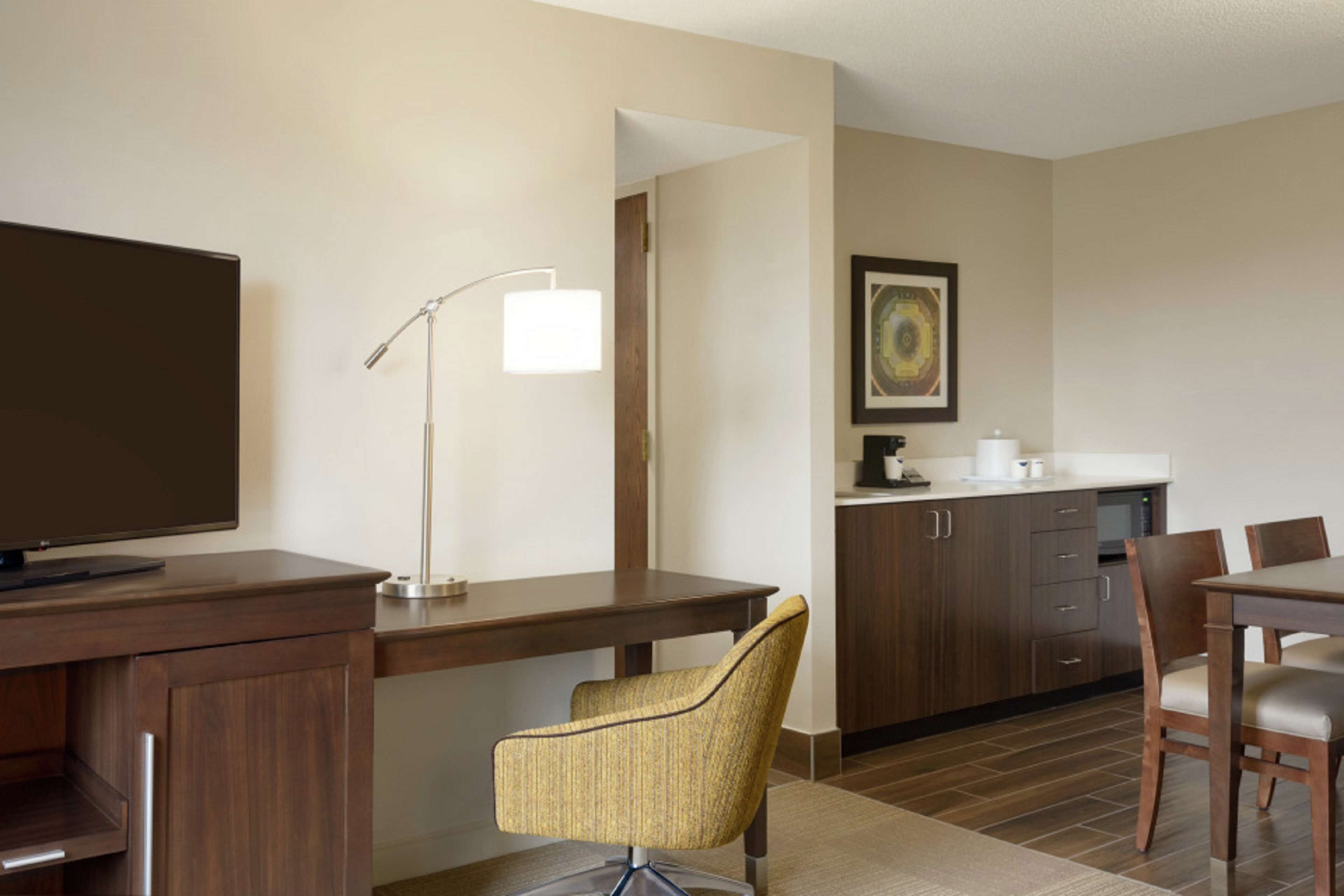 A hotel room with a desk, chair, TV, lamp, and kitchenette.