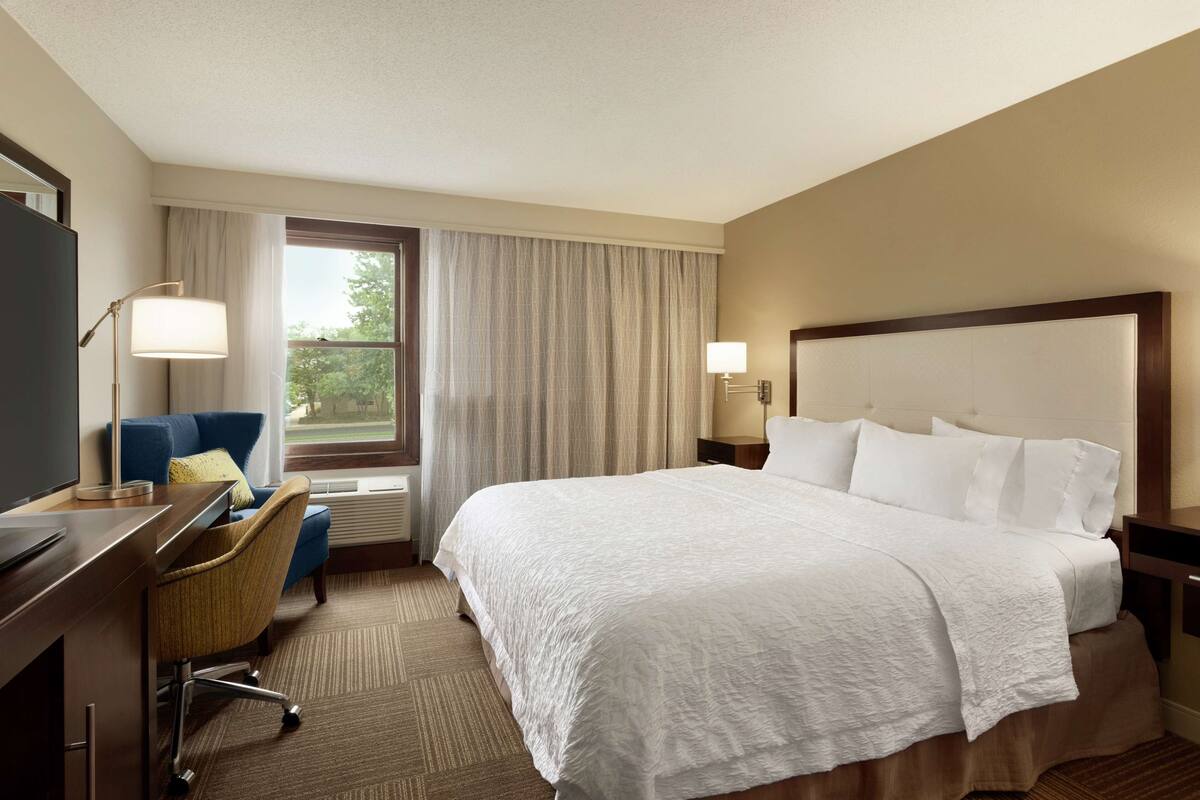 A hotel room with a large bed, a desk with a chair, a TV, and a window with curtains.