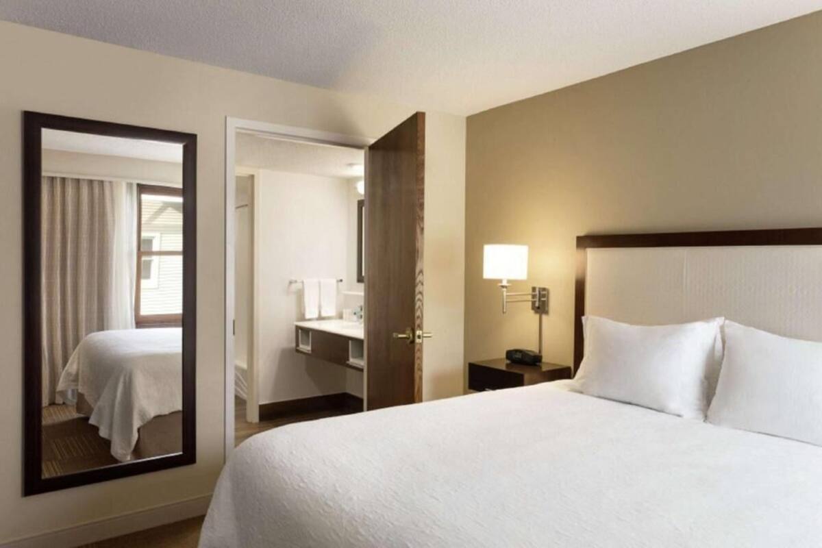 A hotel room with a large bed, a bedside table with a lamp, a mirror, and a door leading to a bathroom.