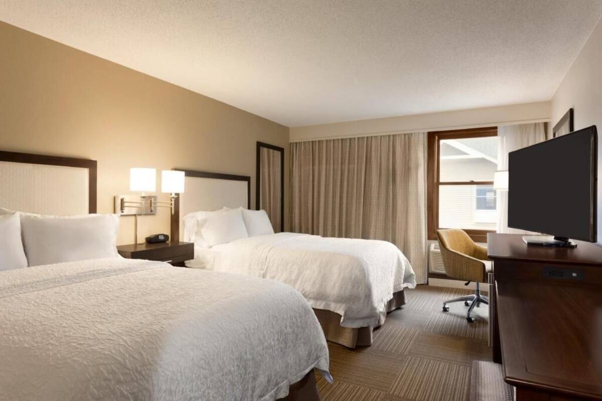 A hotel room with two beds, a desk, a chair, a TV, and a window with curtains.