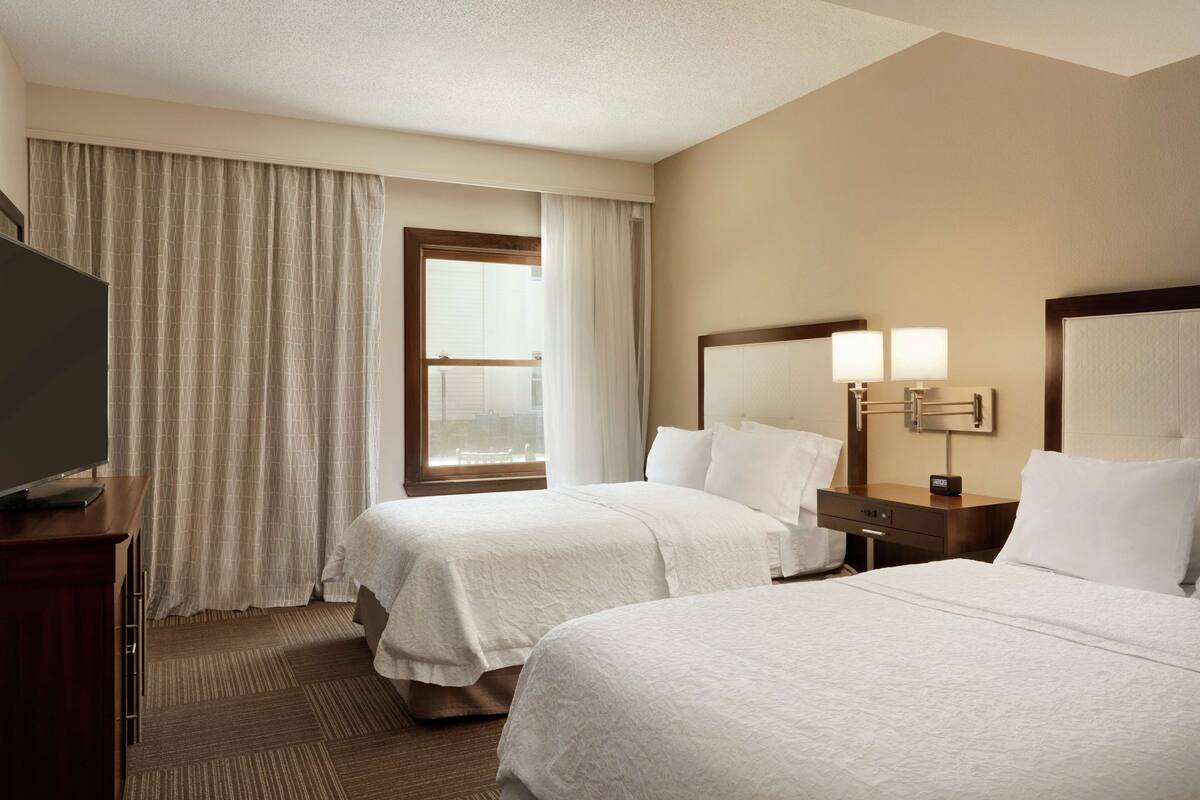 A hotel room with two beds, a television on a desk, a window with curtains, and a nightstand with lamps.