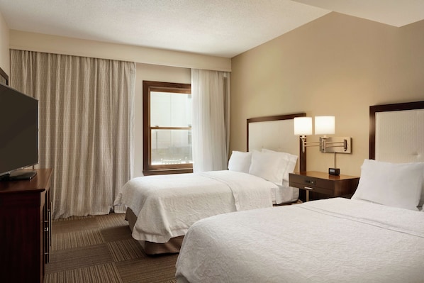 A hotel room with two beds, a television on a desk, a window with curtains, and a nightstand with lamps.