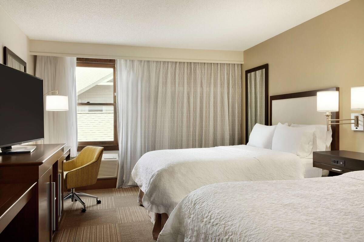 A hotel room with two beds, a desk with a TV, a chair, and a window with curtains.