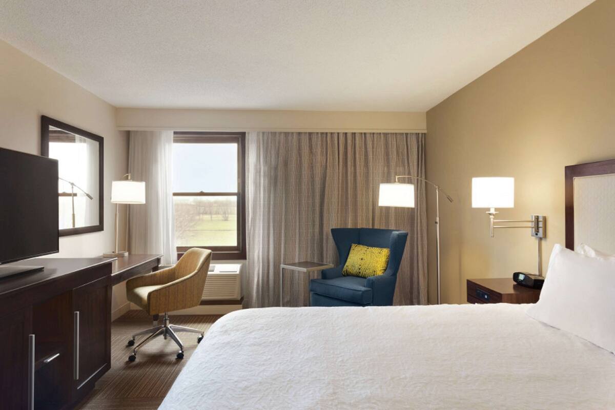 A hotel room with a large bed, a desk, a chair, a TV, a lamp, and a window with curtains.