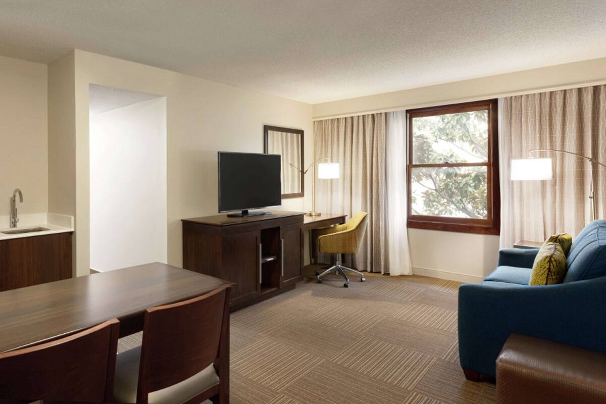 A hotel room with a wooden dining table, a television on a cabinet, a desk with a chair, a sofa, and a large window with curtains.