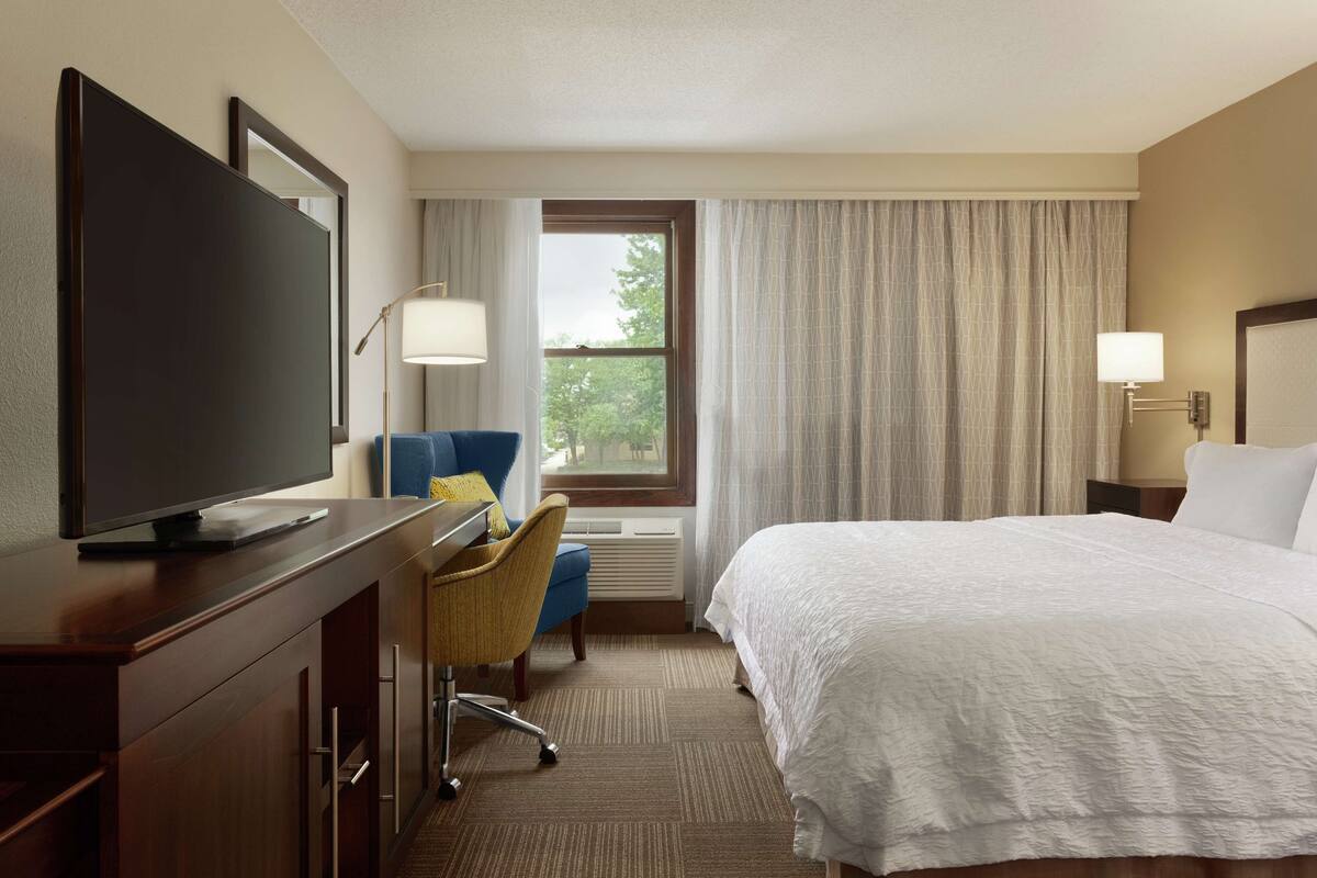 A hotel room with a bed, a desk with a television, a chair, a window with curtains, and a lamp.