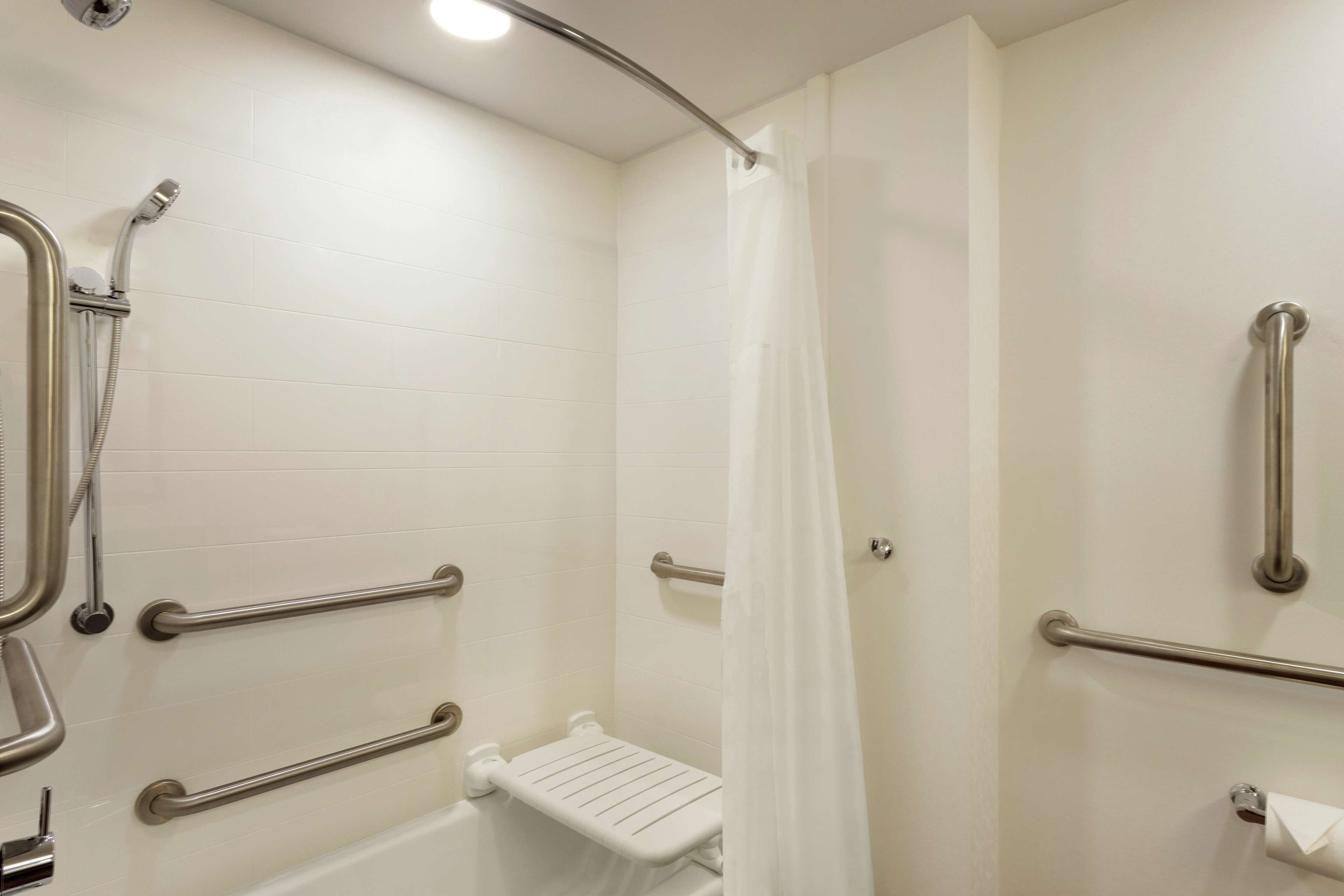 A bathroom with white tiled walls, a shower with a handheld showerhead, and grab bars.