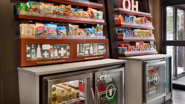 A snack display with various chips and snacks, a refrigerator with beverages, and a 'OH' sign on the wall. - Hampton Inn Youngstown/Boardman (Poland)