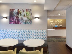 Een hotel lobby met een receptiebalie, een kleurrijk abstract schilderij en zithoeken met ronde tafels.