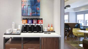 A coffee station with four dispensers labeled 'Light Roast,' 'Dark Roast,' 'Decaf Roast,' and 'Hot Water.' There are stacks of white cups and a small table with a coffee pot and a bottle of lotion.