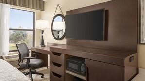 A hotel room with a bed, a desk with a chair, a flat-screen TV, a microwave, a lamp, and a window with a view of trees. - Hampton Inn Youngstown/Boardman (Poland)