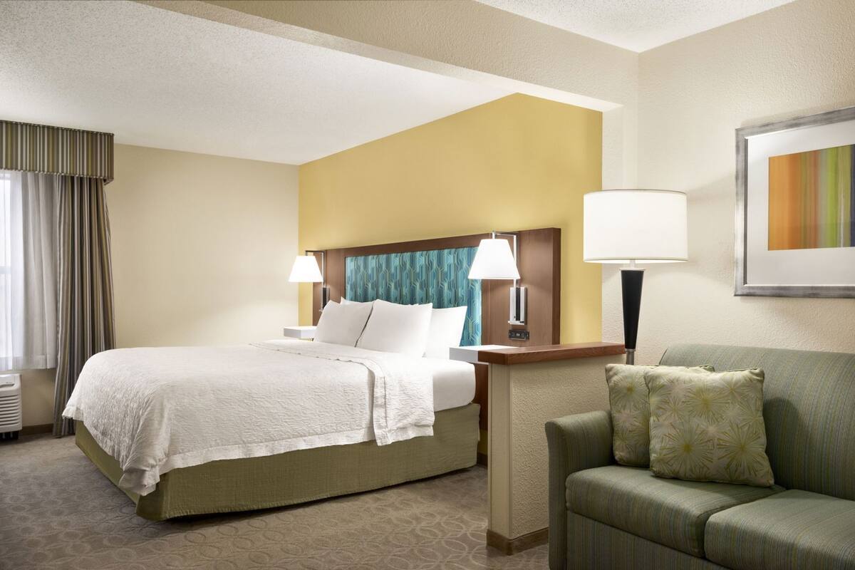 A hotel room with a bed, a sofa, two bedside lamps, a headboard with a patterned design, and a framed picture on the wall.