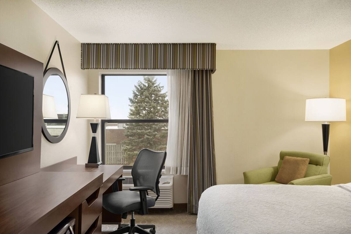 Habitación de hotel con cama, escritorio con silla, televisor, ventana con cortinas y lámpara.
