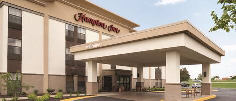 Um Hampton Inn com entrada coberta, uma placa de 'Welcome' e um estacionamento.