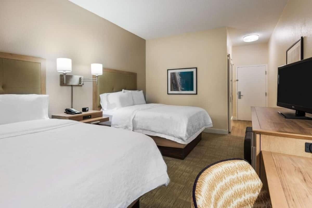 A hotel room with two beds, a desk, a chair, a television, and a wall-mounted lamp.