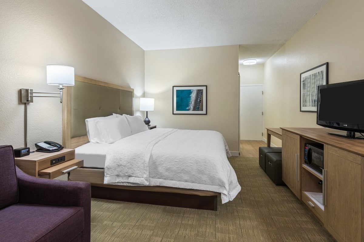 A hotel room with a bed, bedside table with a lamp, a desk with a computer, a TV, and a couch.