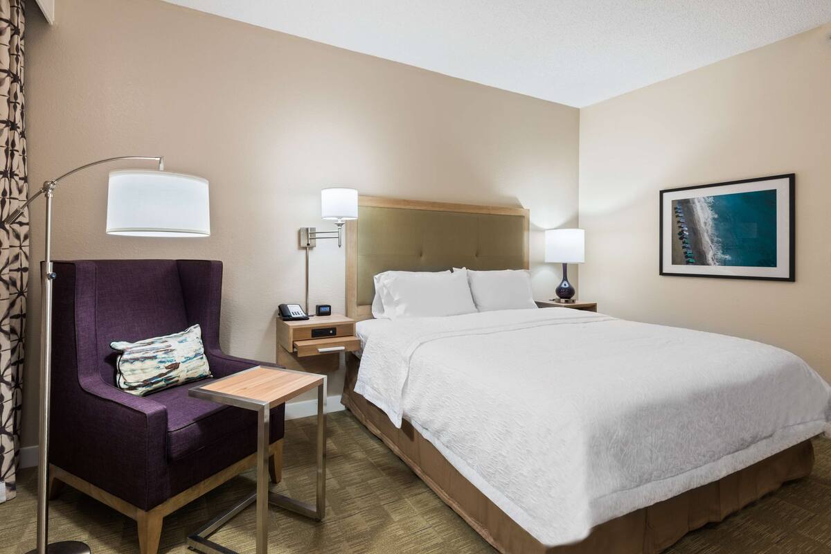 A hotel room with a large bed, a chair with a pillow, a nightstand with a lamp, and a picture on the wall.